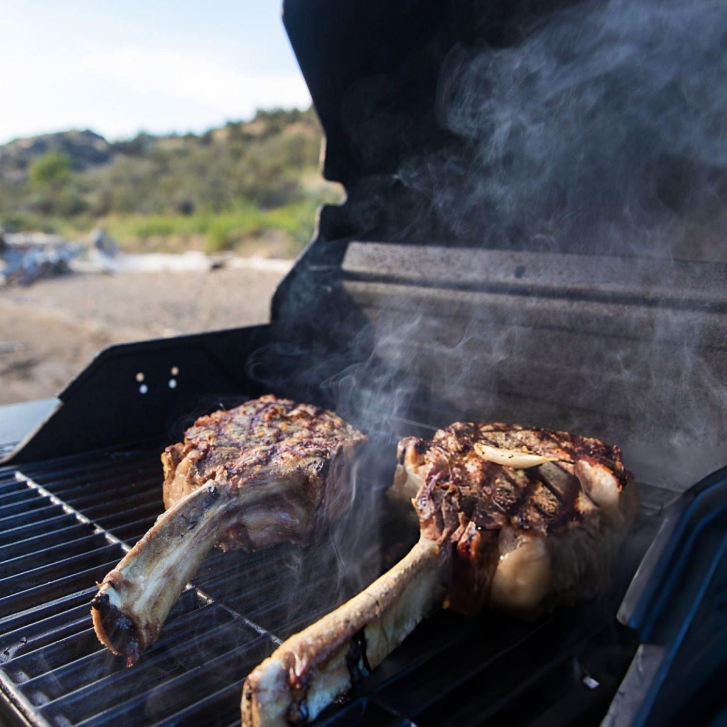 Two bone-in tomahawk steaks cooking on a grill with smoke, outdoors. - 澳洲優質黑安格斯斧頭扒