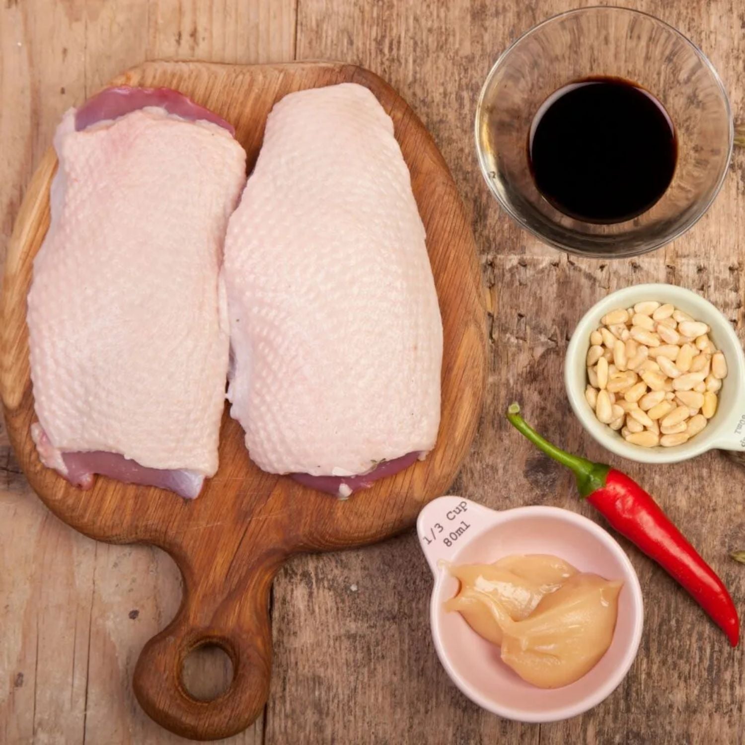 Raw duck breasts on a wooden cutting board with soy sauce, pine nuts, a red chili pepper, and a small bowl of garlic on a rustic wooden surface. - 無激素法國無骨連皮番鴨胸