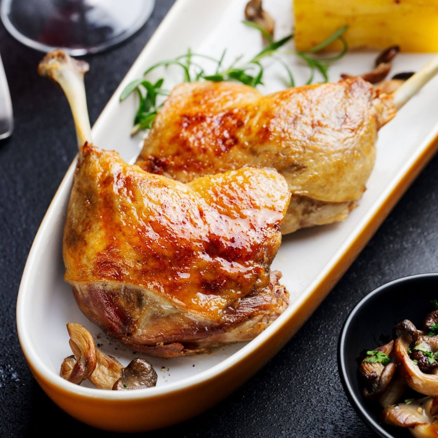 Two roasted duck legs on a white plate with garnishes on a dark surface. - 無激素法國連皮鴨髀