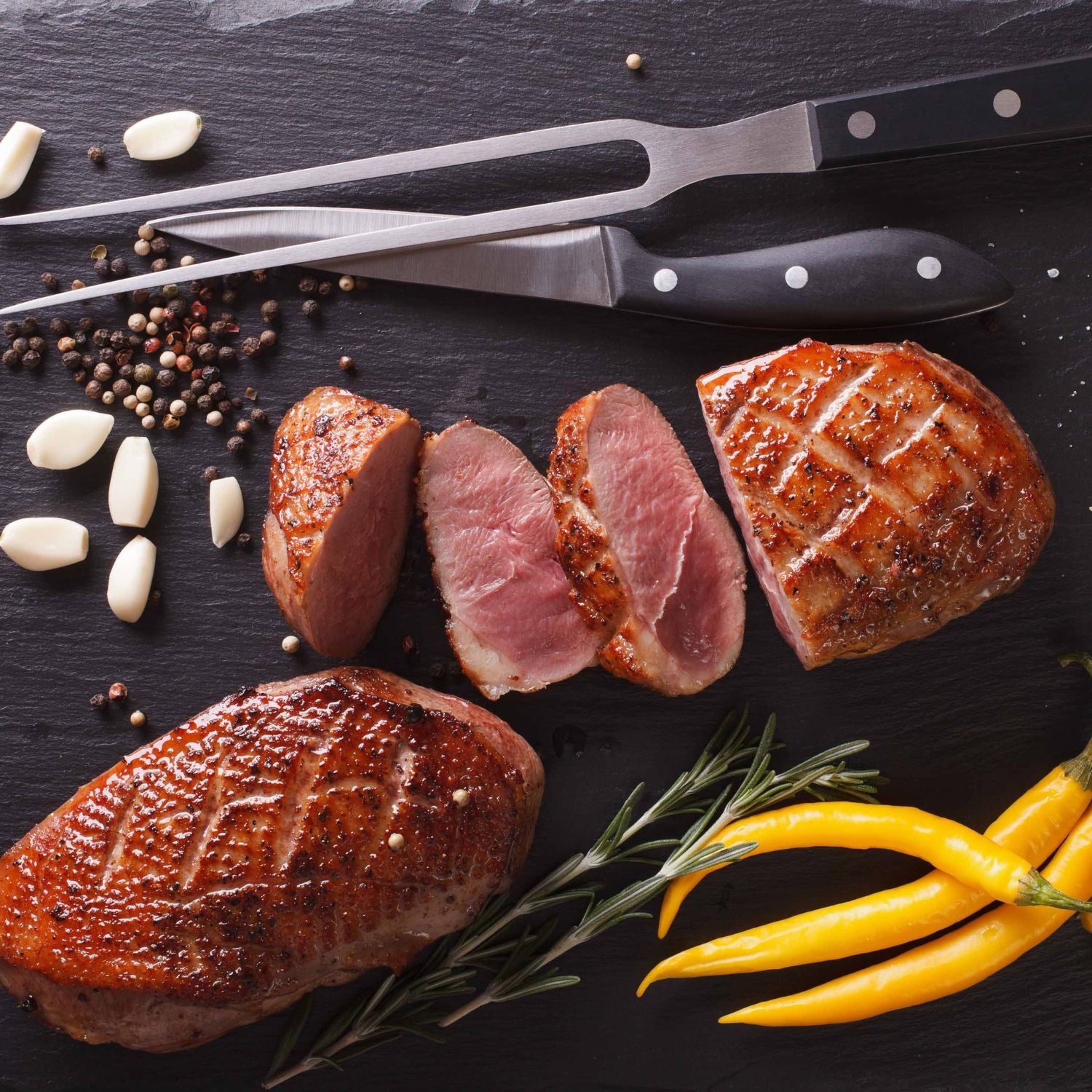 Sliced roasted duck breast with spices, garlic, and peppers on a dark surface. - French Hormone Free Moulard Duck Breast Skin-On Boneless Fillet